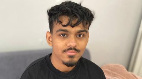 A young man with goatee beard and moustache, wearing a black sweatshirt, sits on a grey sofa in a house. He is looking at the camera.