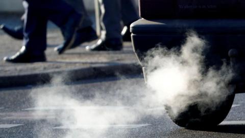 The rear of a car is visible with fumes coming out of the exhaust. Pedestrians are visible in the background.