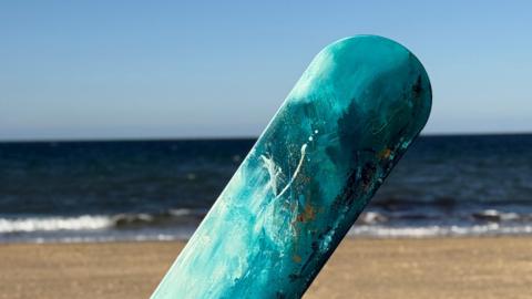 A picture of one of the painted body boards in Cornwall. It is mainly light blue in colour with the beach and sea in the background.
