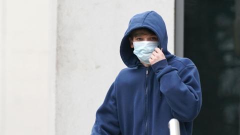 A man wearing a dark blue hoodie and a light blue face mask stood outside a white building.