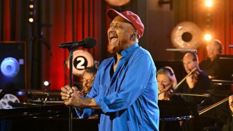 Labi Siffre, wearing a blue shirt and red cap, singing with the BBC Concert Orchestra in Radio 2's Piano Room