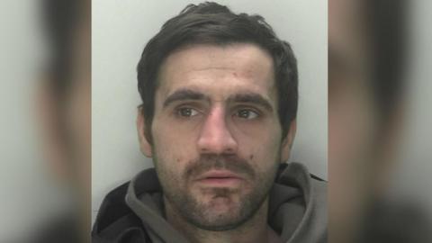 A mugshot of a man in his early 30s, but with quite a weathered face with frown lines and dark spots, who has brown hair. He is looking away from the camera, his mouth ever so slightly agape, and his nose looks as if it has been broken before. He is wearing a grey hoodie, the hood of which is visible, and is standing against a plain, dull white background.