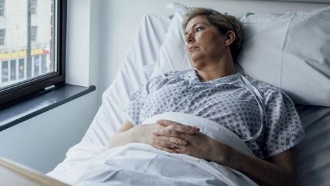 A woman lies in a hospital bed and looks out of a window