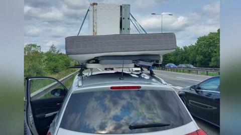A grey car which is parked on the side of the M11 motorway with the door open. Attached to the roof of the vehicle is an outdoor foldable table with a mattress on top and then a white fridge on the top of the pile.