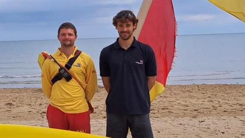 Two men standing on a beach. A red and yellow flag can be seen in the background, as well as the sea and a blue sky. The man on the left is wearing red trousers and a yellow long-sleeve polo top. He is wearing a black strap with a radio connected to it. He has short brown hair and short brown facial hair. The man on the right is wearing black jeans and a navy short-sleeve polo shirt with a logo on. He has short curly brown hair and short brown facial hair. He is also wearing black sunglasses on his head.