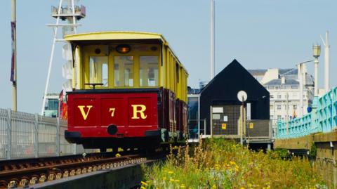 A small red and yellow train runs along a track in an urban setting. 