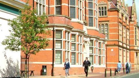 Brick building with more than 18 visible windows. There are people walking on the pavement, a tree, a waste bin, a bench and three black bollards.