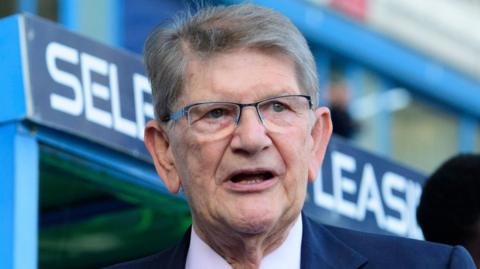 A picture of Sir John Madejski, who has greying hair and glasses and is wearing a suit, in front of a dug out at the SCL Stadium in Reading.