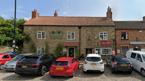 A Google Maps image of the Hansom Restaurant in Bedale, a large stone building that neighbours a pizza takeaway. The building has five large windows with white frames, and a black drain pipe down the middle dividing the two properties and their front doors. Hansom has a tiled awning above their black door, and there are seven cars parked along the street in front of it. The property has a chimney, a tree growing directly next to the restaurant, and greenery is growing up the bricks. There is also a street light off to the left.