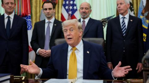 A man in a suit sits at a desk with his hands held out. 