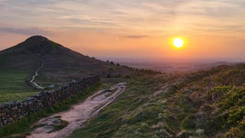 A sunset from the top of Roseberry Topping. A stone wall runs alongside a path running up the peak of the hill in the distance.