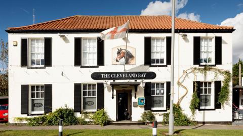 The white exterior of The Cleveland Bay with black shutters open around the windows. A picture of a brown horse can be seen above the main entrance.