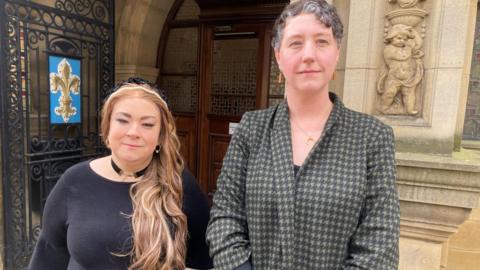 Two women stood outside a council building. The woman on he left has long blonde hair with a headband and is wearing a black top and black choker-style necklace. The woman on the fight has short, dark grey hair and is wearing a grey and black checked jacket. 