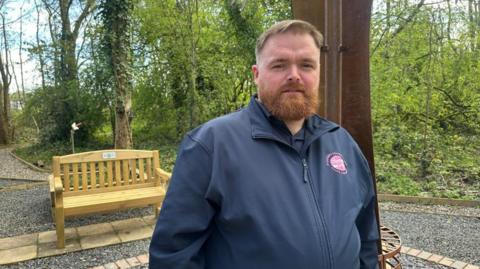A man with short brown hair and a ginger beard in a black top standing in front of a bench in a memorial garden.