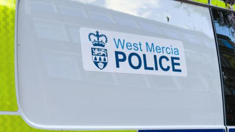 A West Mercia Police sticker on a police van which is predominately white on the panel pictured with yellow high viz stickers around it.