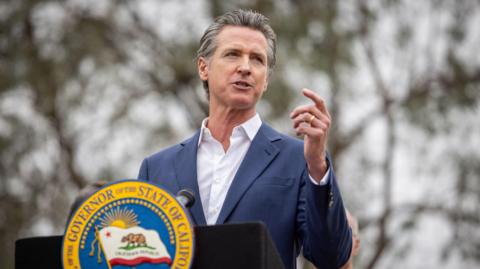 California Governor Gavin Newsom standing at a lectern, speaking at a press conference, with one arm slightly raised