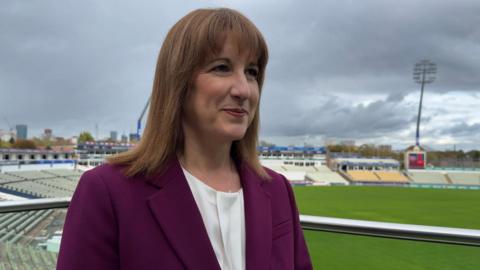 A woman wearing a purple blazer and white blouse stands on a balcony at a stadium. Grass and rows of seating can be seen behind her.