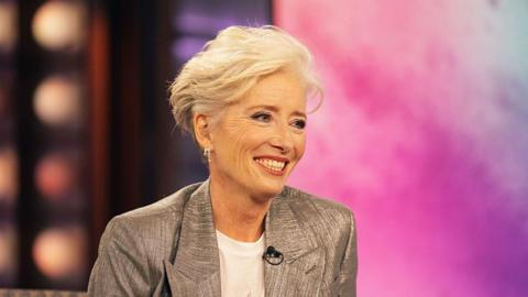 Emma Thompson sat on stage on the Kelly Clarkson show. She is smiling whilst wearing a white t-shirt and silver blazer. 
