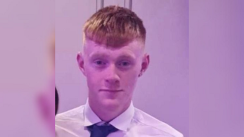 A young man with ginger hair stares at the camera as the picture is taken. He is wearing a white shirt and blue tie. 