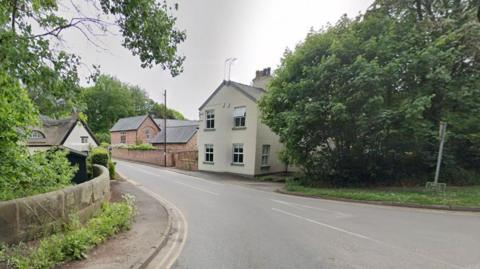 The start of Mill Lane, Mobberley, in Cheshire, showing a property with a thatched roof on the left and on the right some trees then two properties  