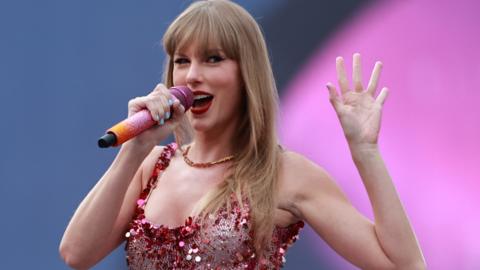 Taylor Swift performing on stage at the Aviva Stadium in Dublin, during the Eras Tour. Taylor is wearing a pink sequined top with a matching coloured microphone in her right hand. She is waving with her left hand and is looking off to her left
