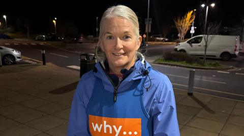 Rachel Clark wearing a blue running jacket underneath a blue vest for the Why cancer charity. She has silvery-blonde hair which is swept back into a low ponytail, and is smiling at the camera. She is standing on a wide pavement beside a road at night.