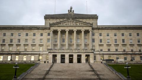 A view of Parliament Buildings from beneath the steps of the Stormont Estate. It is a large, detached, four-storey over basement Neoclassical Portland limestone construction with six pillars on its central portico. The are neat lawns and two black cast-iron standard lamps on either side of the steps.  A statue of Britannia, flanked by two lions, is on top of the building.