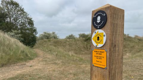 At the forefront of the photo on the right is a post, with three signs on. The top is a black circle, reading 'King Charles III England Coast Path.' The sign underneath is a white circle with a yellow arrow which is pointing to the left, also reading 'King Charles III England Coast Path.' The bottom line reads 'Public Right of Way.' In the background, to the left, is a path surrounded by grass and trees. 