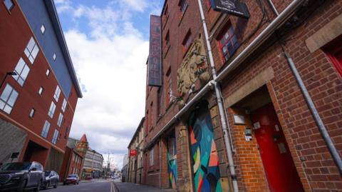 A street with a four-storey building  on one side. There is a black sign outside with the words 'The Leadmill' in red, neon writing. Two doors to the building are painted in multi-colours. 