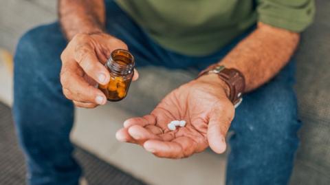 A man in a green t shirt and blue jeans holding some white pills in his hand