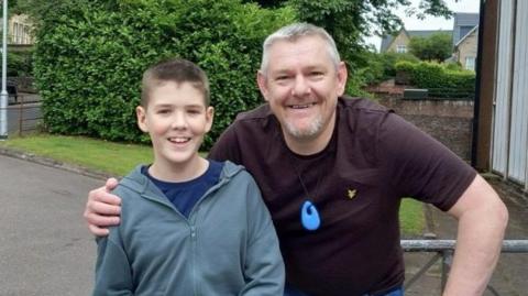 There is a young boy wearing a grey coat and blue pants. He has brown hair. He is being hugged by an older man with grey hair. He is wearing a maroon t-shirt, around his neck is a blue silicone toy. They are stood in front of some trees. They are both smiling. 