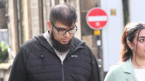 Mohammad Rashid leaving court in a black jacket with glasses on. His face is angled down as he looks up to the camera. 