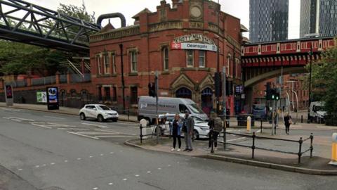 Street view image of the junction of Whitworth Street West and Deansgate,  showing people waiting to cross the road, with the red brick building of Deansgate railway station in the background