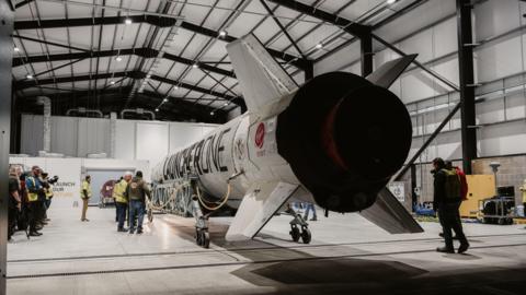 LauncherOne rocket at Spaceport Cornwall, at Cornwall Airport in Newquay. The rocket can be seen from the rear with four fins at the back of the craft. LauncherOne is written in big letter down the side of the rocket. Several people can be seen in the shot getting the rocket ready.