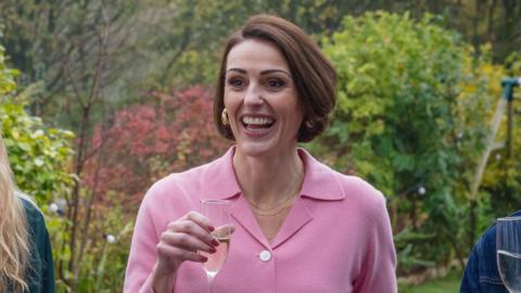 Character Suz laughs in a garden while holding a glass of fizz. She has brown hair and wears a pink shirt