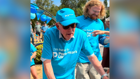 David Treasure in a blue Parkinson's UK t shirt with a Parkinson's UK cap and sunglasses. He has his arms outstretched towards his walker which is out of shot. There are people to the side of him and behind and a balloon arch with Parkinson's UK balloons. 