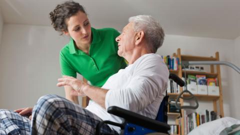 A woman in a green t-shirt is talking to a man in a wheelchair. She has her brown curly hair pulled back in a ponytail. He has grey hair and is wearing checked trousers and a long-sleeved white top. They are probably pyjamas.