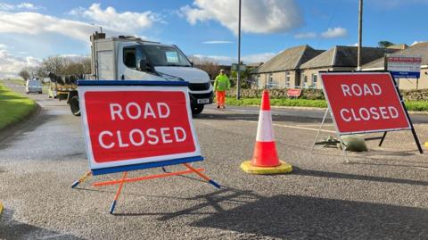 The picture of the scene with a road closed sign at the top of the street. Behind the sign is a vehicle with a police car seen in the background.