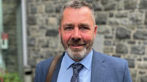Deputy Marc Leadbeater - A man with black and grey hair and a a similar beard. He is wearing a blue suit with a blue shirt and a blue tie. 