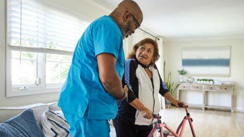  A male care worker is supporting a woman who is using a walking frame 