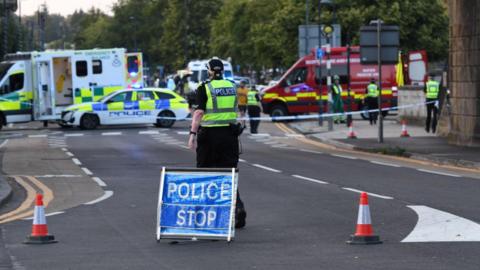 A road blocked off by police with emergency service vehicles in the background