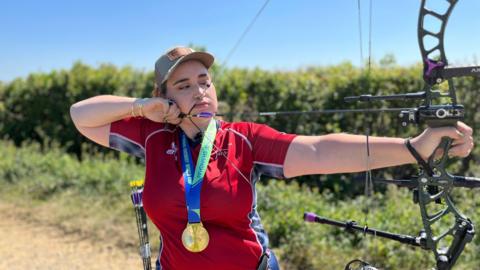 A woman wears a red sporty shirt and a gold medal around her neck, while pulling a bow. She looks very concentrated as if she's about to shoot an arrow. She has a small number of arrows behind her waste ready to use next.