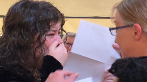 One student on the left with her hand over her face while she cries and is greeted by another person. They have result papers in their hands