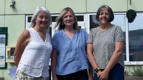 Three middle-aged women stand in front of a pale green building. A sign reading "The Buckland Centre" is on the wall behind them.