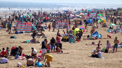 Families on Skegness beach, with crowds of people using windbreakers and beach umbrellas. The sea is in the background.