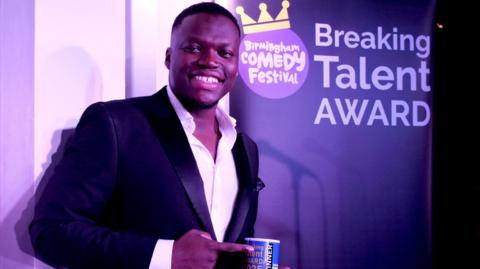 A man with very short black hair is wearing a black suit jacket and white shirt. He is holding a mug that is blur and white and reads "breaking talent award 2025. he is pointing at it with his free hand. Behind him is a large banner that reads "breaking talent award 2025"
