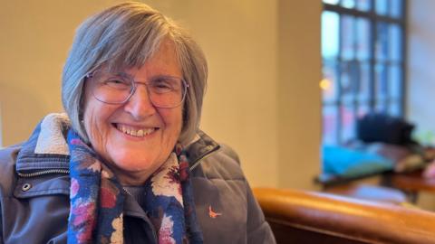 Mary Warren is smiling at the camera while sitting inside the new hub. She wears thin, purple-framed glasses, a purple coat and a pink, blue and purple scarf. She has grey hair which has been styled into a face-framing bob