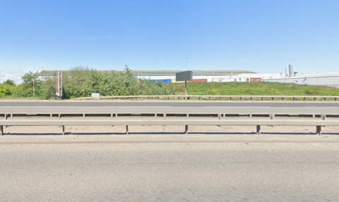 The A66 is a large dual carriageway. It is a sunny day and there is the large grey retail park in the background. There is also a sign in the background and large streetlights. 