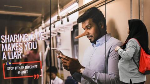 A commuter (R) walks past an advertisement reading 'sharing a lie makes u a liar' at a train station in downtown Kuala Lumpur on March 26, 2018