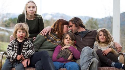Maria and Nik Payne sit on a sofa leaning into each other and touching foreheads, as their four children sit around them